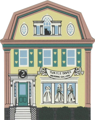 3/4" thick wooden replica of an illustration of a building facade with the sign 'Turtle Doves Wedding Gallery' above a storefront window displaying wedding dresses. The building has a pale yellow face, teal shutters and door, pink trim, and a dark green base, with a black cat silhouette visible in the upper window.