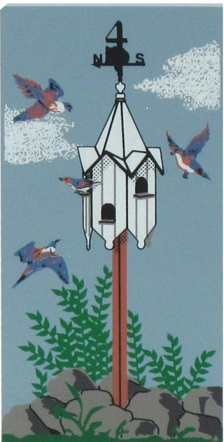 3/4" thick wooden replica of an illustration of a white birdhouse mounted on a brown post, surrounded by flying birds, green plants, rocks, and a blue sky background.