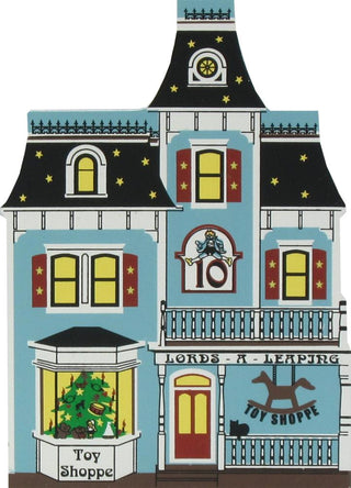 3/4" thick wooden replica of an illustration  of a house with a light blue facade, dark roof, yellow windows, red shutters, and storefront text reading 'Lords A Leaping Toy Shoppe' and 'Toy Shoppe'.