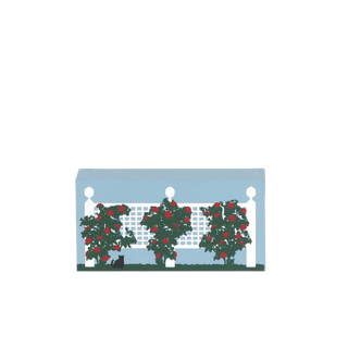 Decorative wooden replica with a white fence, greenery, and red flowers on a light blue background