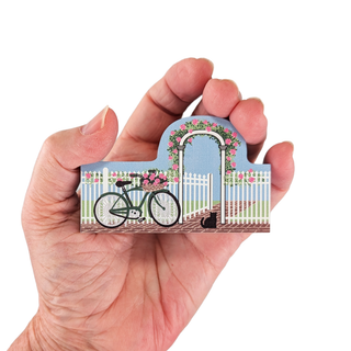 Hand holding a 3/4" thick wooden illustration of a bicycle, flowers, and a cat in front of a white picket fence and floral archway.