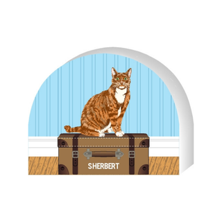 Wooden replica of a cat named Sherbert sitting on a suitcase with a blue wall background.