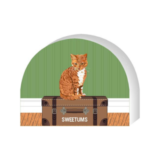 Wooden replica of a  Cat sitting on a suitcase with 'Sweetums' branding against a green wall.