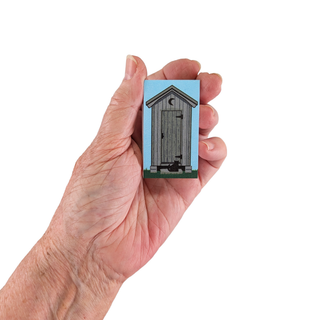 Hand holding a 3/4" thick wooden figurine with a design of a wooden outhouse against a blue background.