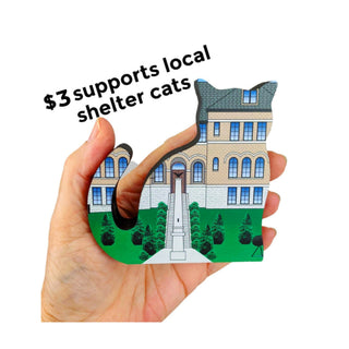 A hand holds a shaped wooden cat-shaped sign featuring an illustration of a shelter cat house with green lawn and black text reading '$3 supports local shelter cats' on a white background.