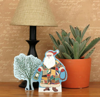 Decorative wooden cutout of a patchwork Santa Claus figure on a table with a lamp and plant in the background.