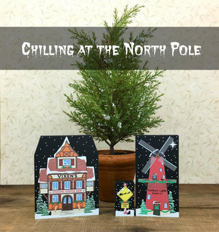 Two Christmas-themed cards with a house and windmill design, placed in front of a small potted tree.