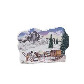3/4" wooden replica with an illustration of a snowy mountain scene with a sled and people, featuring the word 'Alaska'.