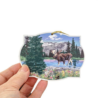 Hand holding an ornament of a moose in a natural landscape with mountains and trees.