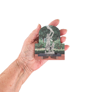 Hand holding a 3/4" wooden replica with an illustration of with a statue and trees on a white background