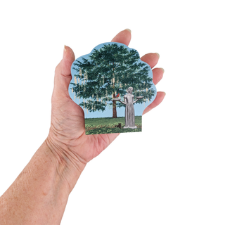 Hand holding a 3/4" wooden replica with an illustration with a scenic design of a statue under a tree.