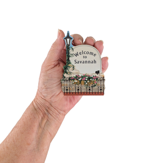 Hand holding a decorative sign with 'Welcome to Savannah' text on a white background