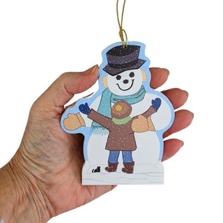 A hand holding a die-cut snowman Christmas ornament featuring a smiling snowman and child in winter clothing, with a gold hanging string against a white background.