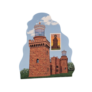 A wooden decorative plaque or sign shaped like a lighthouse and lighthouse museum building, printed with a blue sky, clouds, green grass, and a small postage stamp graphic showing a lighthouse.