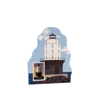 A die-cut wooden plaque or magnet featuring a vintage-style illustration of a white lighthouse on a dark rocky base under a light blue sky, with a small postage stamp graphic of a lighthouse in the lower left corner.