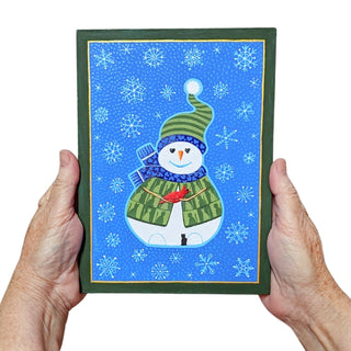 A painting featuring a winter snowman illustration, held by two hands against a plain white background.