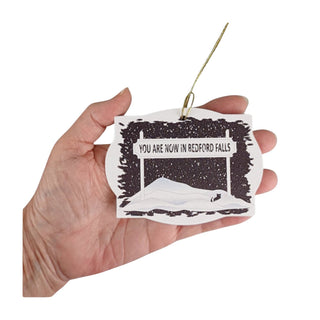 Hand holding a paper ornament with 'You are now in Bedford Falls' text and black and white illustration.