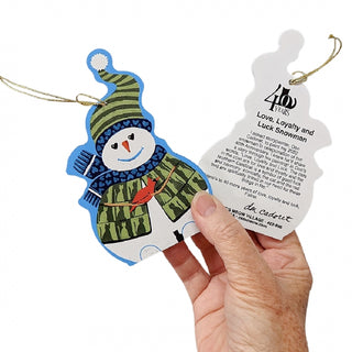 A hand holding a wooden Christmas ornament shaped like a snowman next to a matching silhouette tag with printed text. The ornament is decorated with a green striped hat, blue scarf, and red bird detail, while the tag reads '400 Years Love, Loyalty and Luck Snowman' and shows the 'J.