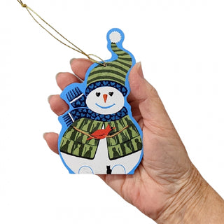 A hand holding a wooden snowman Christmas ornament with a green striped hat and scarf, blue trim, and a small red bird in its hands, on a white background.