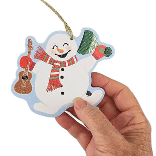 A hand holding a die-cut snowman Christmas ornament with a gold hanging string. The ornament features a white snowman wearing a red and green striped scarf, a green knit hat, and holding a brown guitar, set against a plain white background.