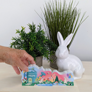 Hand holding a colorful card with a white rabbit and plants in the background