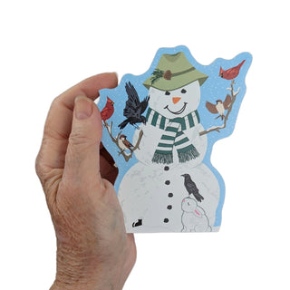 hand holding 3/4" thick wooden replica with an Illustration of  a white snowman with a green striped scarf, green hat, black buttons, birds perched on branches, and a small white bunny on a light blue snowy background.