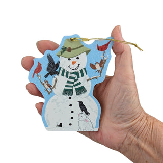 A hand holding a die-cut Christmas ornament shaped like a snowman with birds and a rabbit, featuring a blue border and a gold hanging string, against a plain white background.