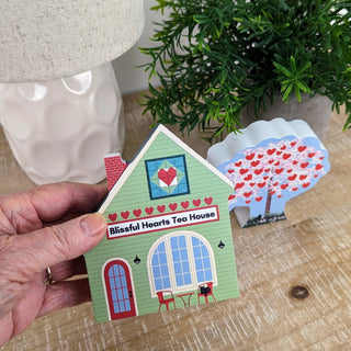 Hand holding a 3/4" thick wooden illustration of a tea house with a heart design and 'Blissful Hearts Tea House' sign.