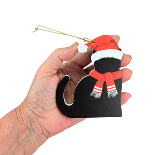 A hand holding a black cat Christmas ornament wearing a red Santa hat and red and white striped scarf, with a gold hanging string, against a plain white background.