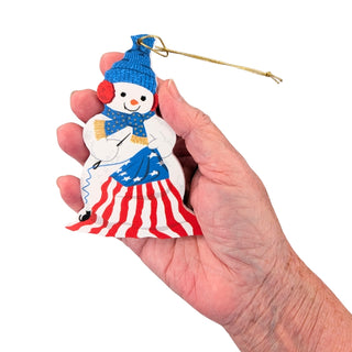 A hand holding a small decorative snowman ornament with a blue hat and scarf, red and white striped skirt, and a gold hanging loop, shown against a plain white background.