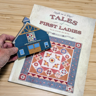 Hand holding a 3/4" thick wooden illustration of a blue barn with quilt design with 'Lady of the White House' text next to a book titled 'Tales of First Ladies' on a wooden surface.