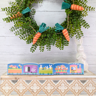 Decorative Easter train set with colorful cars on a wooden shelf, surrounded by greenery and a white rabbit figurine.