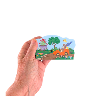 Hand holding a small illustrated wooden replica with a cartoon scene of a car, house, and trees on a white background.