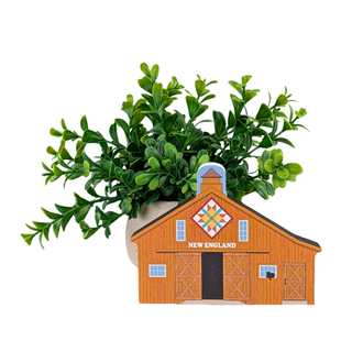 An artificial green boxwood plant in a light beige pot, styled behind a decorative wooden New England barn ornament on a solid white background.