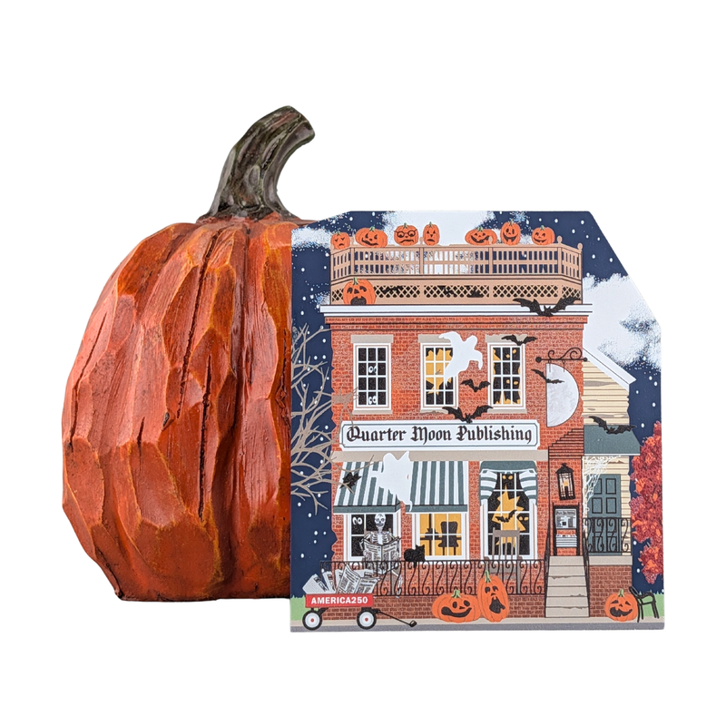 A carved orange pumpkin sits beside a Wooden 3/4" replica with an  illustrated card featuring a Halloween storefront with the text 'Quarter Moon Publishing' and 'American Nightmare.' The background is plain white.