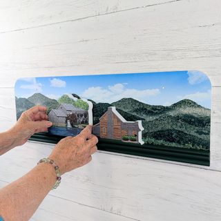 Person holding a wood figurine with a scenic drawing of a cabin and mountains on a white wooden surface.