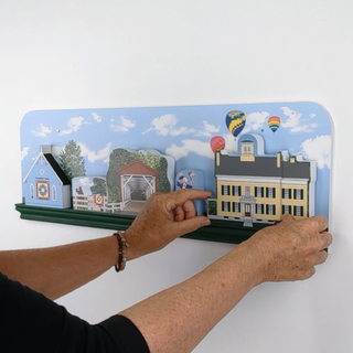 Hand interacting with a colorful 3D wall shelf depicting buildings and nature on a white background.