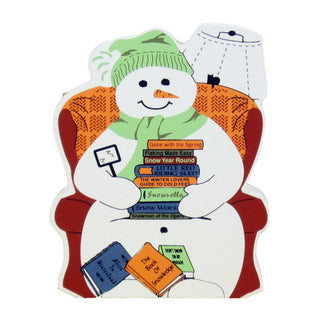 3/4" thick wooden replica with an Illustration of a snowman sitting in an orange armchair, wearing a green knit hat and scarf, with stacked books and a small black cat figure.