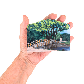 Hand holding a 3/4" wooden replica with an illustration with a scenic illustration of a bridge and trees.