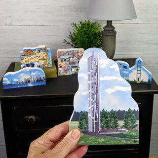 Hand holding a 3/4" thick wooden cutout of a building illustration in front of a display of similar buildings on a table.