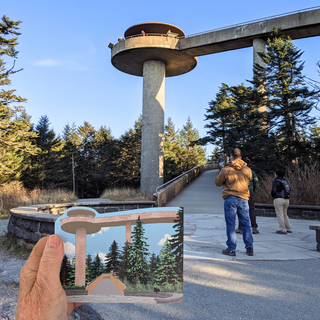 A hand holds a small wooden replica of Clingman's Dome at Great Smoky Mountain National Park, along with a walkway, trees, and several people under a blue sky.