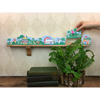 Decorative shelf with cartoon designs, a hand holding additional cartoon pieces, books, and a plant on a wooden surface.