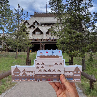 A hand holds a small wooden replica of a lodge building in front of a real outdoor scene with trees, grass, a path, and a large stone lodge under a cloudy blue sky.