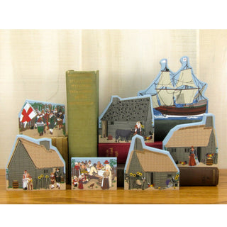 Set of miniature models of a ship, houses, and people on a wooden surface with a book in the background.