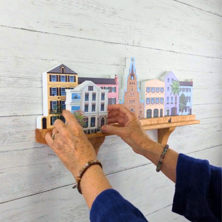 Hands arranging colorful miniature buildings on a wooden shelf against a light wooden background