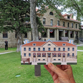 A hand holds a wooden replica of Albright Visitor Center building in front of a real outdoor scene with grass, trees, and the actual stone building in the background.