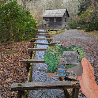A hand holds a small printed or painted landscape scene featuring a weathered wooden cabin, a narrow wooden bridge over flowing water, and leaf-covered ground. The larger real-world scene in the background shows the same environment in full, with trees, fallen leaves, and the cabin visible.