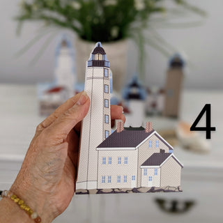 A hand holds a small paper model of a lighthouse and house, with a blurred background showing similar miniature models and a plant. A large black number 4 is overlaid on the right side.