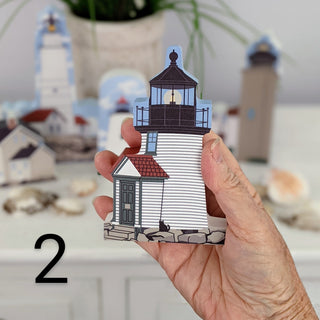 A hand holds a small illustrated lighthouse-shaped item, with a blurred background showing similar lighthouse decor and seashells on a tabletop. A black number 2 appears in the lower left.