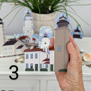 A hand holds a small illustrated paper lighthouse and house cutout in front of a larger assembled lighthouse scene on a tabletop, with a black number 3 overlay in the lower left.